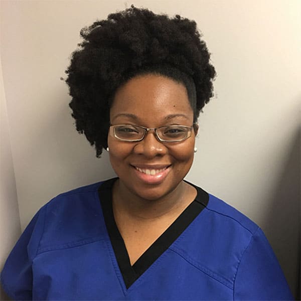 Smiling woman in blue scrubs with glasses standing against a light wall She has natural curly hair pulled back from her face Smiling woman in blue scrubs with glasses, standing against a light wall. She has natural, curly hair pulled back from her face.
